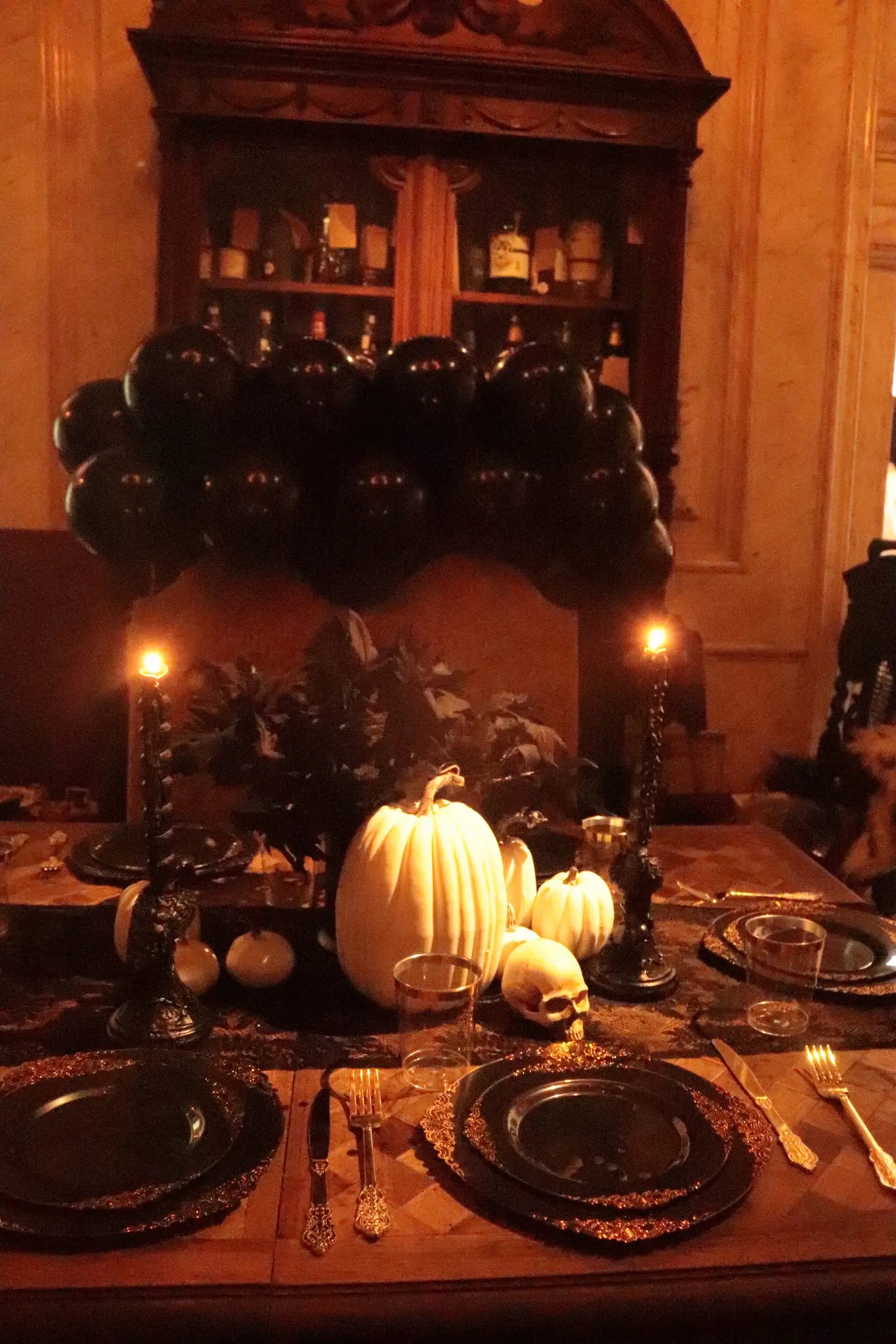 Black and gold Halloween party table with white pumpkins, skull decor, black candles, lace runner and black balloon backdrop