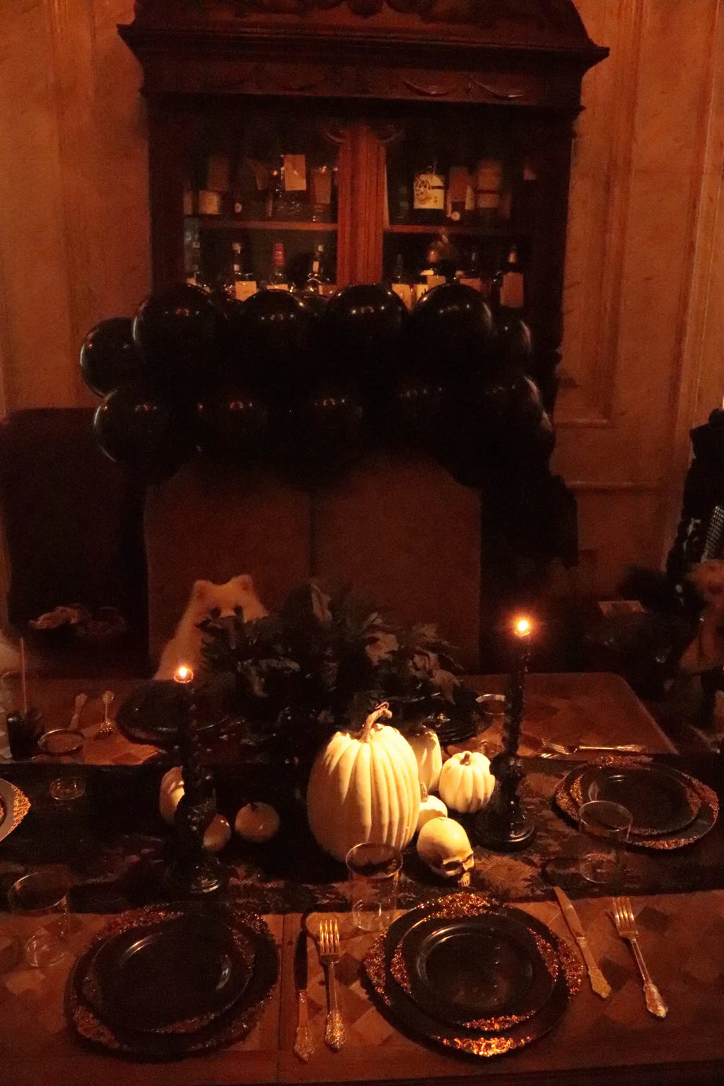 Gothic Halloween dinner party table with black balloon garland, white pumpkins, skull decoration, candle holders and black gold plates
